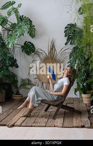 Junge, entspannte, verträumte Frau, die einen Papierventilator verwendet, während sie sich im schönen, erfrischenden Hausgarten entspannt Stockfoto