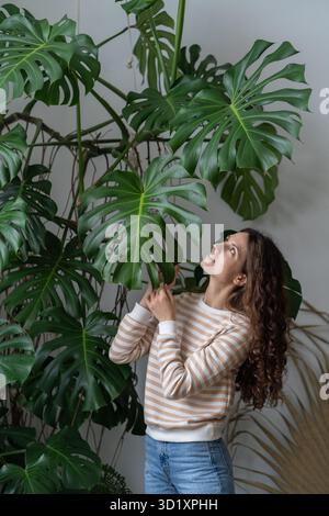 Junge lächelnde Frau berührt das Monstera-Pflanzenblatt und genießt die Gartenarbeit und plantcare Stockfoto