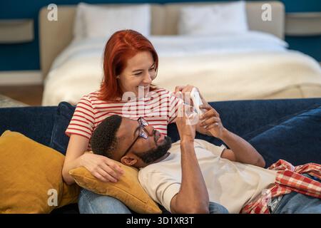 Ein junges, vielfältiges Paar, das sein Smartphone benutzt, wählt das Hotel für den Urlaub, während es sich zu Hause auf dem Sofa ausruht Stockfoto