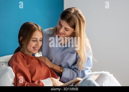 lächelnde mutter und Teenager-Tochter lesen Buch, lernen Verse zusammen, während sie zu Hause im Bett sitzen Stockfoto