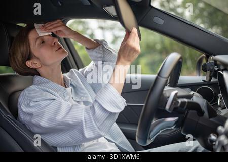 Frau fährt Auto mit defekter Klimaanlage bei heißem Sommerwetter, wischt Schweiß-Papierserviette ab. Stockfoto