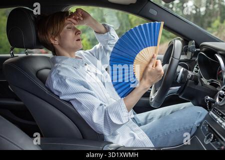 Eine müde, erschöpfte Frau mittleren Alters, die mit einem blauen Fächer wedelt, leidet an Steifigkeit im Auto. Stockfoto