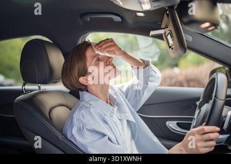 Frau fährt Auto mit defekter Klimaanlage bei heißem Sommerwetter, wischt Schweiß-Papierserviette ab. Stockfoto