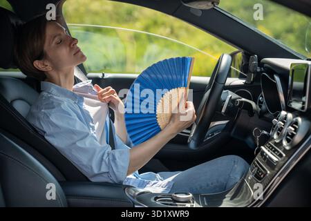 Überhitzte, erschöpfte Fahrerin mittleren Alters mit Handventilator, die an Hitze im Auto leidet. Heißes Wetter Stockfoto