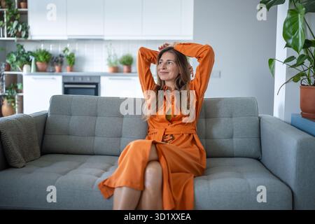 Die lächelnde, glückliche Frau liegt auf der Couch, die Hände liegen hinter dem Kopf und genießen die Zeit allein im Wohnzimmer Stockfoto