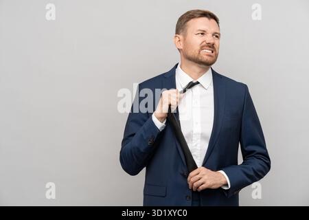 Ein müder Geschäftsmann legt Krawatten im offiziellen Anzug ab, mit einem unzufriedenen Gesicht auf grauem Hintergrund. Stockfoto