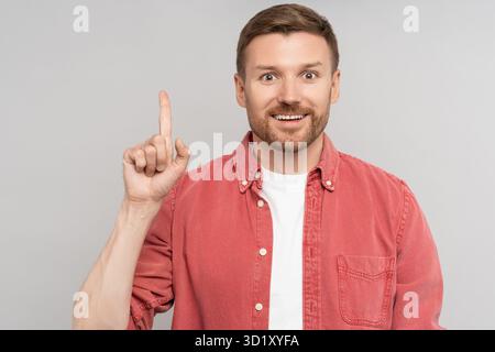 Glücklicher überraschter unrasierter Mann, der den Zeigefinger nach oben auf den freien Platz im Hintergrund zeigt und die Kamera betrachtet. Stockfoto
