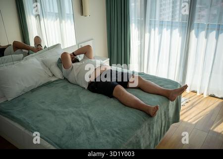 Fette übergroße Männer leiden an Hitze. Fauler, übergewichtiger Mann liegt auf dem Bett und fühlt sich müde im Zimmer Stockfoto
