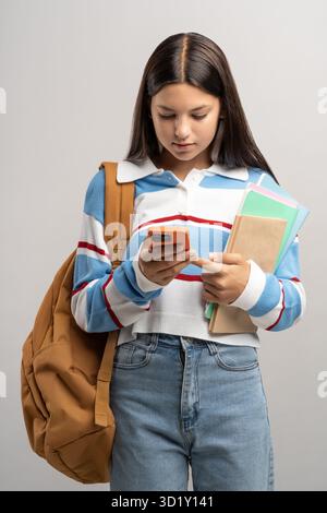 Interessierte Schülerin im Teenageralter mit Rucksack, die auf den Smartphone-Bildschirm auf grauem Hintergrund blickt. Stockfoto