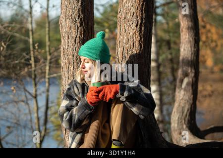 Wehmütige Frau, die auf dem Boden sitzt, nachdem sie in Kiefernwäldern gelaufen ist, heißen Tee getrunken und sich in Sonnenstrahlen erwärmt hat Stockfoto