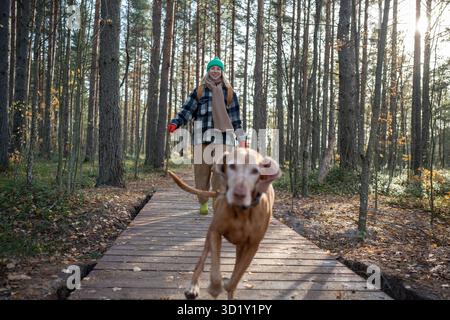Glückliche, lächelnde Tierliebhaberin läuft auf einem Holzweg im skandinavischen Kiefernwald, während sie mit dem Hund spielt Stockfoto