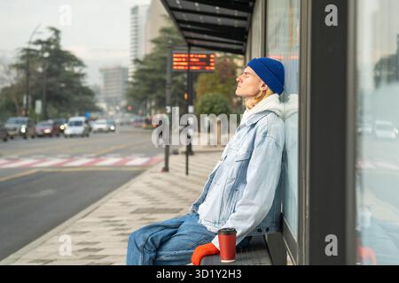 Müder Mann mit geschlossenen Augen, der auf den Transport wartete und morgens nach der Nachtarbeit an der Bushaltestelle saß. Stockfoto