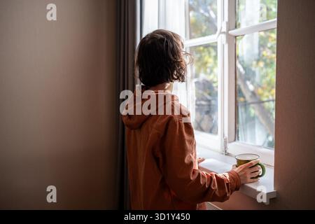 Frau, die auf das Fenster schaut und Tee Kaffee in der Küche zu Hause trinkt, Rückansicht. Stockfoto