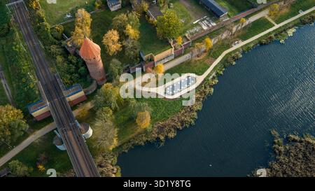 Aus der Vogelperspektive auf die Burg Malbork in Nordpolen, roter Backsteinturm, Mauern am Fluss Nogat, Eisenbahnbrücke, Uferpromenade, Herbstbäume, langes Gold Stockfoto