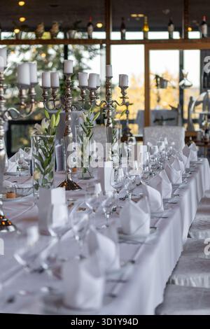 Ein langer Esstisch ist wunderschön mit weißen Tischdecken, Kandelabern und floralen Mittelstücken in einem stilvollen Restaurant eingerichtet. Das warme LIG Stockfoto