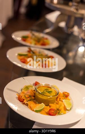 Bei einer Gourmet-Veranstaltung werden drei elegante Lachsplatten kunstvoll auf einem Tisch angeordnet. Jeder Teller ist mit bunten Beilagen und einer lebendigen Sauce, Creati, versehen Stockfoto