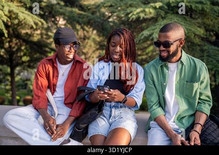 Lebhafter afroamerikanischer Freund sitzt im Park, Telefon in der Hand. Frau teilt trendige Social-Media-Beiträge Stockfoto