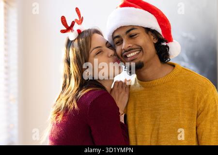 Ein Paar, das zu Hause in der küssenden Wange am Fenster hängt, trägt Weihnachtsmütze, Geweih-Stirnband, Strickpullover Stockfoto