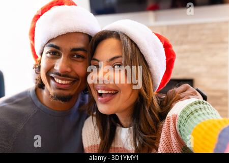 Ein Paar, das sich beugt und lächelt, macht Selfie im Wohnzimmer mit roten und weißen Weihnachtsmützen Stockfoto