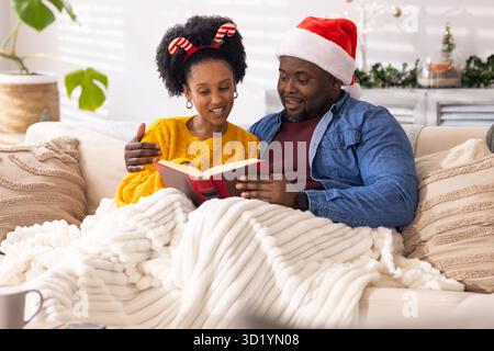 Diverse Paare lesen rotes Buch unter Strickdecke auf dem Sofa im Wohnzimmer mit Weihnachtsmütze Stockfoto