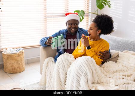 Afroamerikanisches Paar kuschelt unter Decke, Mann bietet grünes Geschenk und Frau reagiert zu Hause Stockfoto