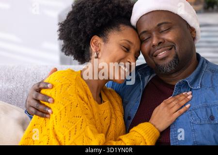 Diverse Paare, die sich auf einem hellgrauen Sofa im Wohnzimmer umarmen, Weihnachtsmütze und Ring tragen Stockfoto
