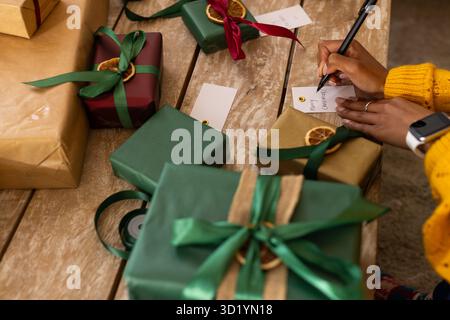 Hände in gelbem Pullover schreiben Geschenkanhänger mit schwarzem Stift auf den Tisch mit Rippenboxen Stockfoto