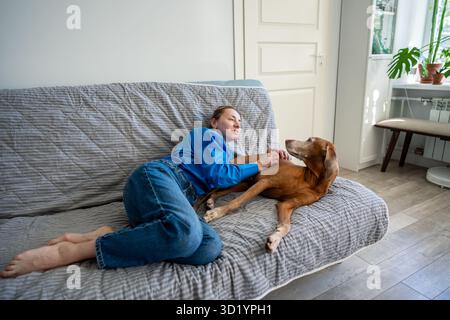 Eine Frau, die auf der Couch sitzt und zu Hause einen Hund umschließt. Entspanntes Wochenende, Komfort für Haustiere, Hundebindung Stockfoto