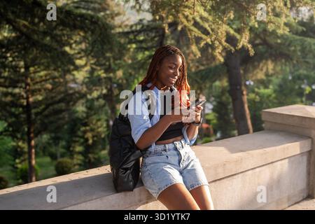 Lächelnde afroamerikanische Studententexte am Telefon im Park, genießen Sie die Einsamkeit, während Sie auf Freunde warten Stockfoto