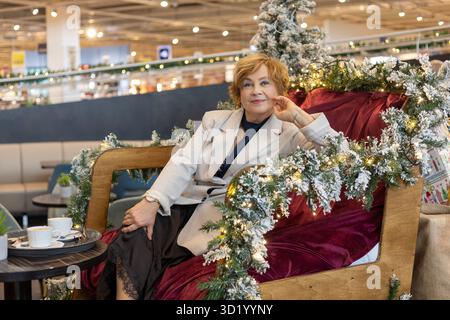 Eine Frau in eleganter Kleidung entspannt sich auf einem luxuriösen Stuhl umgeben von festlicher Dekoration in einem lebhaften Einkaufszentrum und genießt einen ruhigen Moment Stockfoto