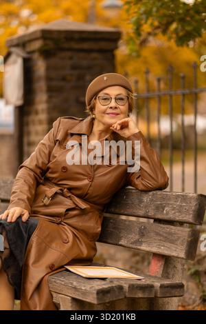 Eine stilvolle Frau genießt die Ruhe eines Parks, während sie auf einer Holzbank sitzt. Die Szene ist mit leuchtenden Herbstfarben wie Orange und Gelb gefüllt Stockfoto