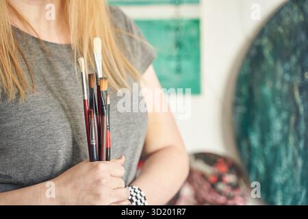 Kaukasische Künstlerin, die Pinsel im Atelier hält, mit Kunstwerken im Hintergrund. Stockfoto