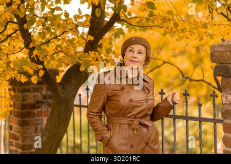 In einer ruhigen Parklandschaft genießt eine ältere Frau in einem stilvollen braunen Mantel die Schönheit des Herbstes. Leuchtend gelbe Blätter sorgen für einen warmen Hintergrund Stockfoto
