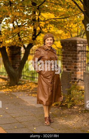 Eine stilvolle Frau steht selbstbewusst auf einem Weg in einem Park, trägt einen braunen Ledermantel und eine passende Baskenmütze, umgeben von leuchtenden Herbstblättern Stockfoto