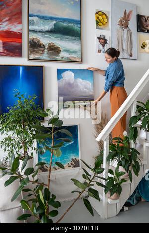 Kreativer Workshop mit Kunstausstellung an Wänden, Künstlerin fügt neue Gemälde zur Galeriewand hinzu Stockfoto