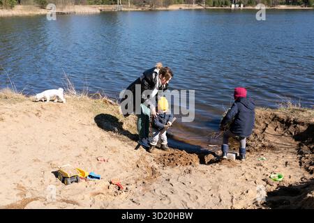 Kinder spielen im Sand am See mit einem Erwachsenen, der an einem sonnigen Frühlingstag unter Aufsicht steht Stockfoto