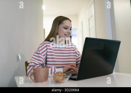 Junge Frau, die tagsüber am Laptop arbeitet und Snacks in gemütlicher Innenumgebung genießt Stockfoto