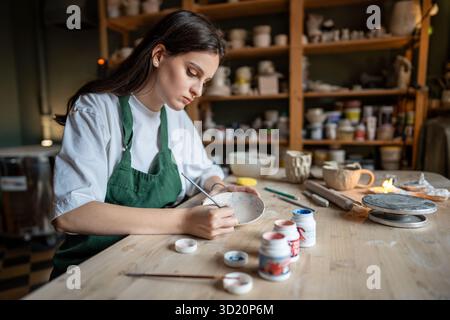 Concentrated female ceramist decorating clay tableware with paints in pottery school. Small business Stockfoto
