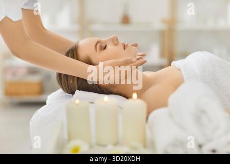 Friedliche junge Frau, die auf einem Massagetisch liegt und im Spa-Center entspannende Gesichtsmassagen genießt. Stockfoto