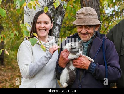 Lächelnde junge Frau und ihr älterer Großvater halten an einem Herbsttag draußen eine grau-weiße Katze, umgeben von Birken und gelben Blättern. Stockfoto