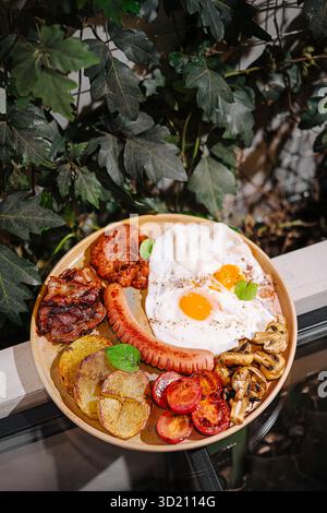 Köstliche Frühstücksplatte mit Eiern, Speck, Würstchen und Gemüse, die im Freien inmitten üppiger Vegetation serviert werden Stockfoto