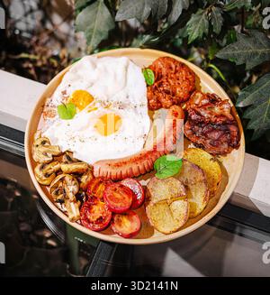 Köstliche Frühstücksplatte mit Eiern, Speck, Würstchen und Gemüse, die im Freien inmitten üppiger Vegetation serviert werden Stockfoto