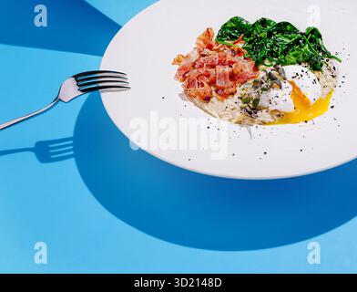 Köstlicher Frühstücksteller mit pochiertem Ei, Gemüse und knusprigem Speck auf einem leuchtend blauen Hintergrund Stockfoto
