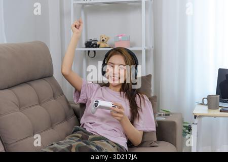 Die junge Frau, die zu Hause glücklich ist, genießt Spielaktivitäten, trägt Kopfhörer und feiert den Sieg mit erhobenem Arm, der den Videospielcontroller auf bequeme Weise spielt Stockfoto