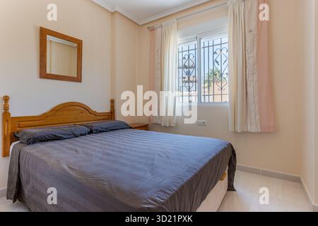 Gemütliches, in neutralen Farben gehaltenes Schlafzimmer mit Holzkopfteil, Spiegel und Blick auf das sonnige Fenster Stockfoto