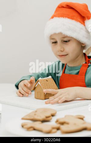 Ein süßer Junge an Silvester und Weihnachten macht in einem Weihnachtshut ein Ingwerhaus in der Küche. Stockfoto