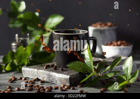 Kaffee spritzt aus einem Kaffeebecher auf einem dunklen Hintergrund, umgeben von gerösteten Kaffeebohnen und Zweigen mit grünen Blättern Stockfoto