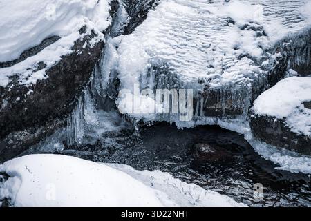Der Fluss Jizerka fließt durch eine kühle Winterszene, umgeben von schneebedeckten Felsen und Eiszapfen. Das kalte Wasser bildet einen schönen Kontrast zur eisigen Landschaft. Stockfoto