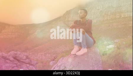 Sitzende Afroamerikanerin im Pullover auf Felsbrocken auf einem Hügel, die in Richtung Sonnenuntergang blickt, Kopierraum Stockfoto
