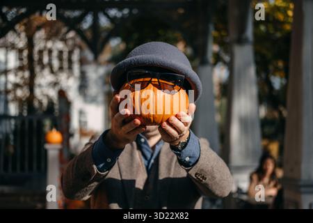 Ein Mann, der einen kleinen Kürbis mit Sonnenbrille und Wollmütze vor seinem Gesicht hält. Gesunder Halloween-Humor, gemütliche Herbststimmung, unbeschwert Stockfoto